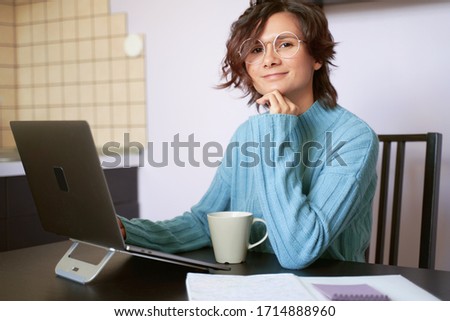 Portrait of young girl looks at camera, leans on chin, sitting in quarantine during pandemic. Beautiful brunette Bob hair wears blue sweater. coffee mug, paper, Notepad, free space for inserting text.