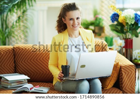smiling modern 40 years old woman in jeans and yellow jacket in the modern living room in sunny day using digital educational content on a laptop.