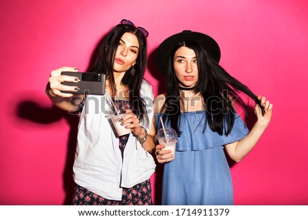 Two caucasian brunette hipster woman in casual stylish outfit having fun making selfie on their phone. They standing on a bright pink background. Cheerful, happy emotions