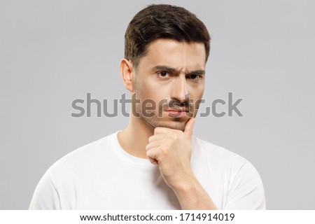 Close-up of young man in white t-shirt, holding his chin, looking at camera with expression of suspicion, mistrusting and doubting, isolated on gray background Royalty-Free Stock Photo #1714914019