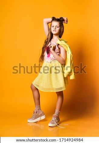 Full length portrait of a cute little teen girl in a stylish skirt and jacket clothes looking at the camera and smiling against a yellow studio wall. Children's fashion concept