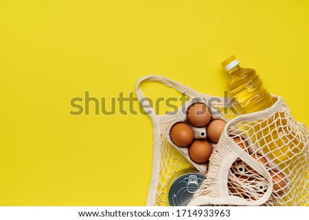 Eggs, pasta, canned food, oil in a string bag on yellow background. Copy space, flat lay. Crisis food stock for coronavirus quarantine isolation period. Food delivery, donation.