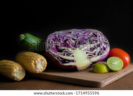 Still life, vegetables on cutting board in a chiaroscuro ambient or low key light, dark food, cabbage, corn, lemon tomato, and zucchini, in black background.