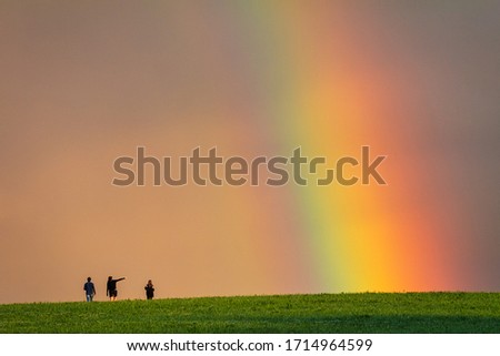 Landscape right after a rain, colorful rainbow over a grassy meadow with young peoples on a walk. 