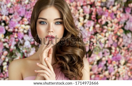 Brunette spring girl with long  and   shiny wavy hair .  Beautiful  model woman with curly hairstyle ,   background  wall of flowers . Skin Care, beauty and  spa