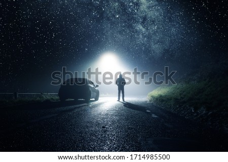 A lone car, parked on the side of the road with a hooded figure, on a spooky country road. With a bright light and stars at night. Royalty-Free Stock Photo #1714985500