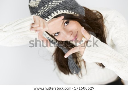 Charming young woman in warm winter clothes, making a photo frame with her hands while looking up and smiling, isolated on grey background.