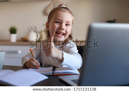 Happy little girl pupil study online using laptop at home, smiling small child show thumb up recommend class or lesson.