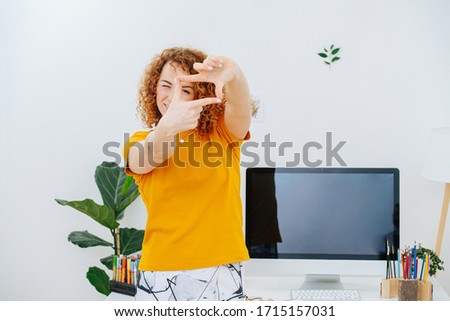 Happy woman artist with gorgeous curly red hair standing in front of her work desk making finger frame, looking through it