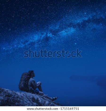 Man hiker with backpack sitting on a cliff above the sea and enjoy the Milky Way in a starry night sky.
