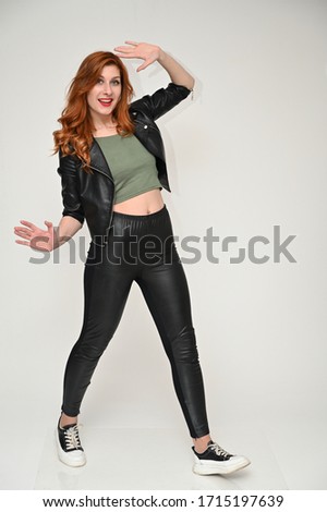 A full-length model with excellent makeup poses in a black jacket and trousers in the studio. Portrait on a white background of a pretty red-haired young woman.
