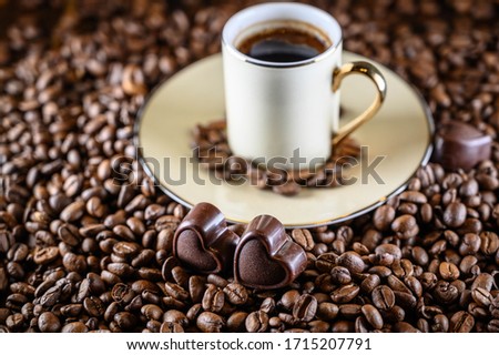 Coffee beans wallpaper. Dark brown roasted coffee bean background with mini heart-shaped chocolate and a cup of dark coffee. Focus on chocolate. Valentine's day celebration idea concept. Side view.