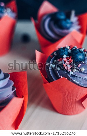 partial close-up view of several chocolate muffins with violet cream in a red wrapper decorated with two blueberries and pastry topping in light pink colors on a wooden tray