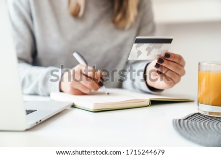 Cropped photo of young woman in eyeglasses making notes in planner while holding credit card at living room