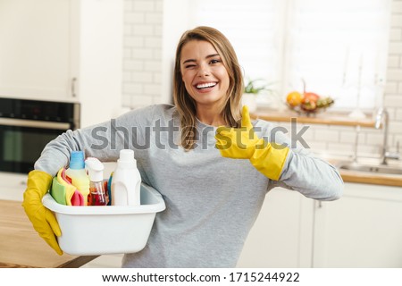 Photo of smiling young woman housewife in gloves holding cleanser bottles and showing thumb up at modern kitchen Royalty-Free Stock Photo #1715244922