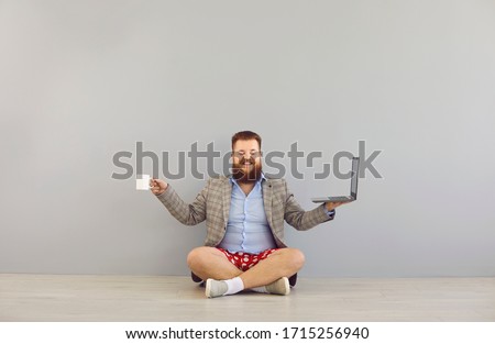 Funny fat bearded man in a jacket and red shorts with a laptop on a gray background.Freelancer online distance work concept.