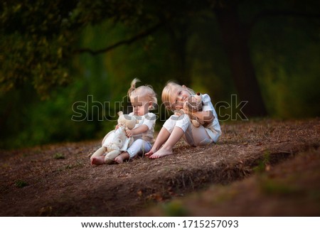 Two blond girls in white summer clothes with tails sitting on a summer glade holding teddy bears