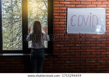The inscription on the wall is the word COVID. A young woman is standing near a window in a room with a brick wall. Self-isolation of a house from a coronovirus