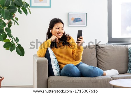 people and leisure concept - happy smiling asian young woman in wireless earphones sitting on sofa and listening to music on smartphone at home