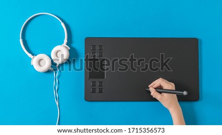 Hand with a pencil on a graphics tablet and white headphones on a blue background. 