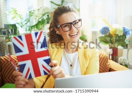 happy trendy student woman in jeans and yellow jacket with laptop showing UK flag notebook in the modern house in sunny day. Royalty-Free Stock Photo #1715390944