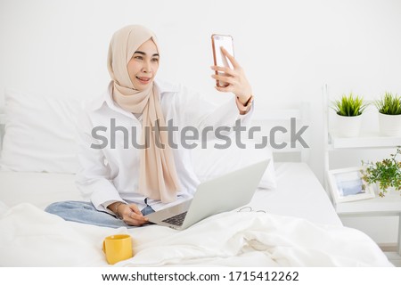 Young pretty muslim woman in white hijab cheerful girl having video-call and photo selfie in white bedroom and working with laptop.
