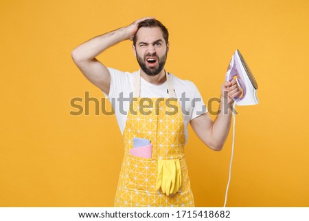 Preoccupied puzzled young man househusband in apron hold in hands iron while doing housework isolated on yellow background studio portrait. Housekeeping concept. Mock up copy space. Put hand on head