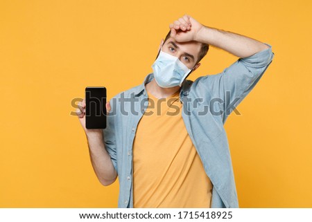 Tired young man in sterile face mask isolated on yellow background. Epidemic pandemic coronavirus 2019-ncov sars covid-19 flu virus concept. Hold mobile phone with blank empty screen put hand on head