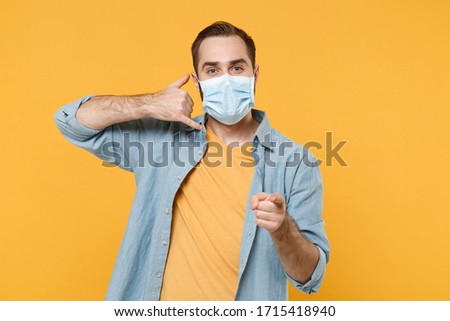Young man in sterile face mask isolated on yellow background. Epidemic pandemic coronavirus 2019-ncov sars covid-19 flu virus concept. Doing phone gesture says call me pointing index finger on camera