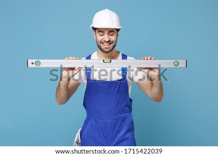 Smiling young man in coveralls protective helmet hardhat hold building bubble spirit level isolated on blue wall background. Instruments accessories for renovation apartment room. Repair home concept