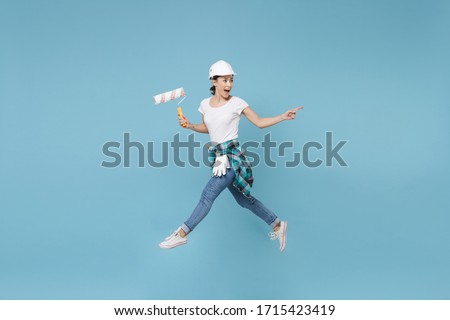Side view excited woman in helmet hardhat jump with paint roller isolated on blue background. Instruments accessories for renovation apartment room. Repair home concept. Pointing index finger aside Royalty-Free Stock Photo #1715423419