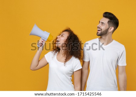 Cheerful couple two friends european guy african american girl in white t-shirts posing isolated on yellow background. People lifestyle concept. Mock up copy space. Scream in megaphone, looking aside