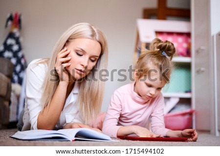 Young mother reads a book at home lying on the floor at home with her daughter, the child watches cartoons on the tablet and plays, happy family, love, family concept.
