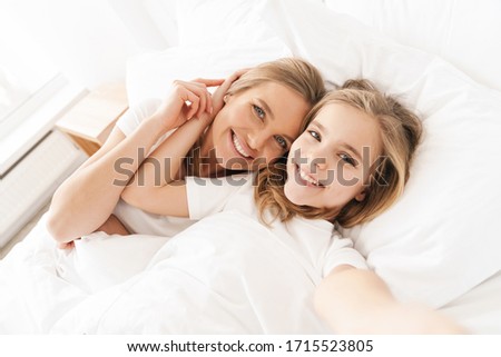 Photo of blonde cheerful mother with daughter smiling and taking selfie while lying in white bad