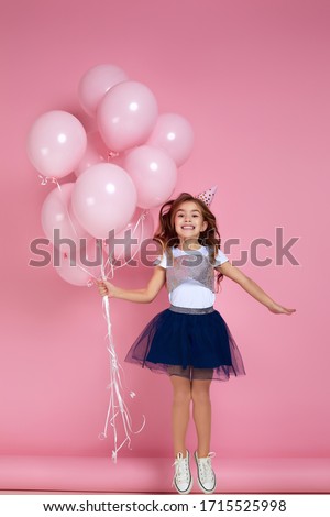 Beautiful happy little child girl in dress and birthday hat jumping with pastel pink air balloons isolated on pink background. birthday party.
