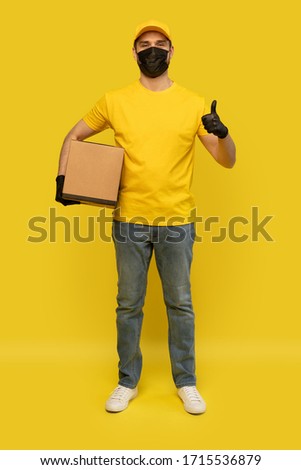 Young delivery man in yellow t-shirt, mask, gloves with box isolated over yellow background. Service quarantine pandemic coronavirus virus 2019-ncov concept