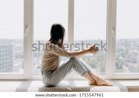 Young woman looking through the window with a city view, sitting on a windowsill, drinking coffee or tea in the morning. Royalty-Free Stock Photo #1715547961