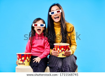 Surprised mother and her little daughter in 3D glasses with popcorn watching movie over blue background. Wow concept                             