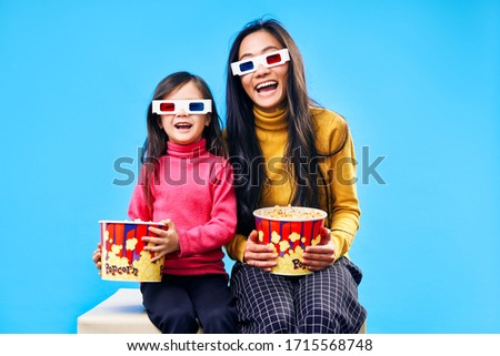 Happy smiling mother and her little daughter in 3D glasses with popcorn watching movie over blue background