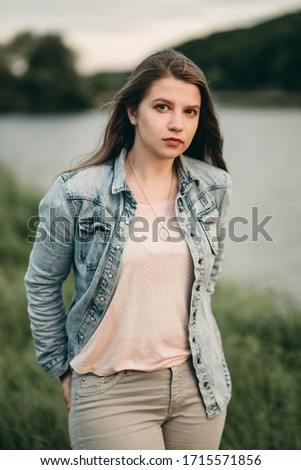 Young white beautiful sad woman in denim, jeans clothes stands on nature in summer near lake at sunset. 