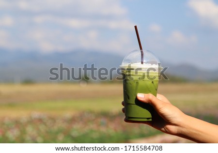 Closeup of woman's hand showing  a cup of cold green tea smoothie with natural background in bright sunny day.