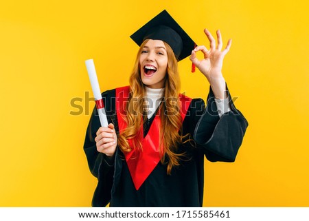 An attractive graduate shows the OK gesture with a diploma in her hands on a yellow background. Concept of the graduation ceremony