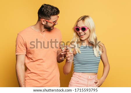 happy young couple in sunglasses clinking cocktail glasses on yellow background