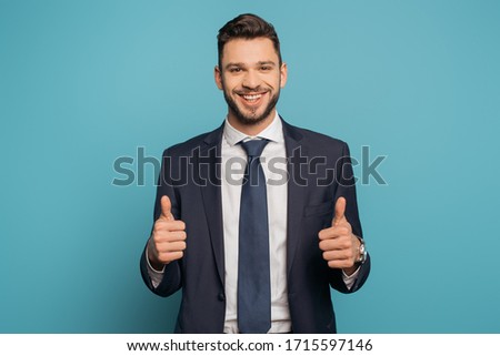 happy businessman showing thumbs up while looking at camera on blue background