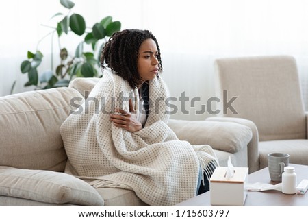 Sick african american woman sitting on sofa covered with blanket, freezing shivering due to high temperature, suffering from coronavirus at home Royalty-Free Stock Photo #1715603797