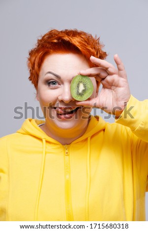 Studio portrait of redhead woman in yellow hoodie covering eye with kiwi fruit on plain background. Healthy eating concept