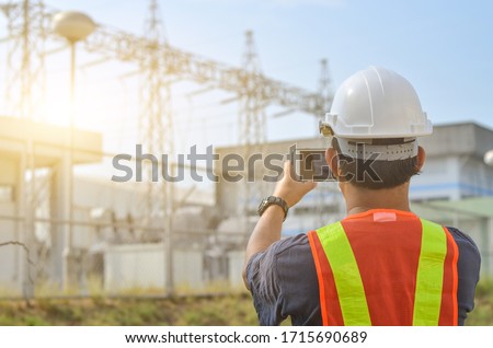 Engineers use mobile phones to photograph high-voltage electricity poles against sub-station background.