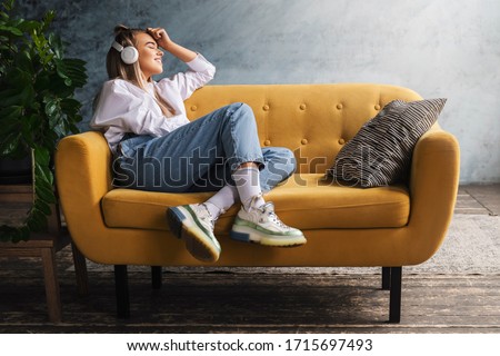 Beautiful blonde woman in white headphones sits on a yellow sofa and enjoys listening to music, podcast. Royalty-Free Stock Photo #1715697493