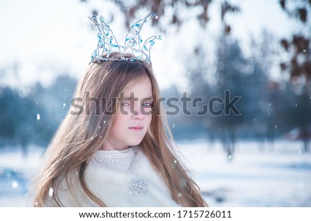 Portrait of a little girl in white fur coat and beaded crown standing on blurry winter snowfall background. 