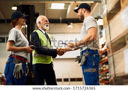 Low angle view of happy senior manager handshaking with a worker while visiting industrial warehouse. 